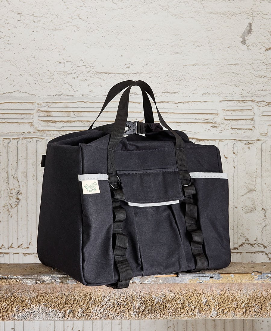 A Black duffel bag with two carry straps, a front flap pocket with hook and loop closure, and additional slip pockets. It is placed against a textured background. Garner and Tow brand