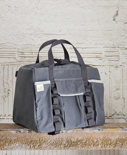A gray duffel bag with two carry straps, a front flap pocket with hook and loop closure, and additional slip pockets. It is placed against a textured background. Garner and Tow Duffel Slate