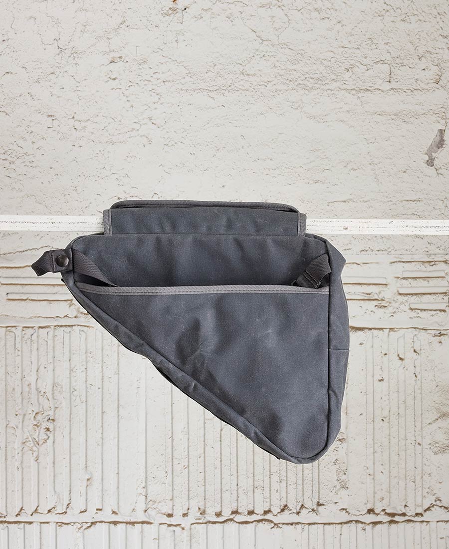 A grey bike frame bag made of waxed canvas, with snap closures and an adjustable shoulder strap, displayed against a textured background.