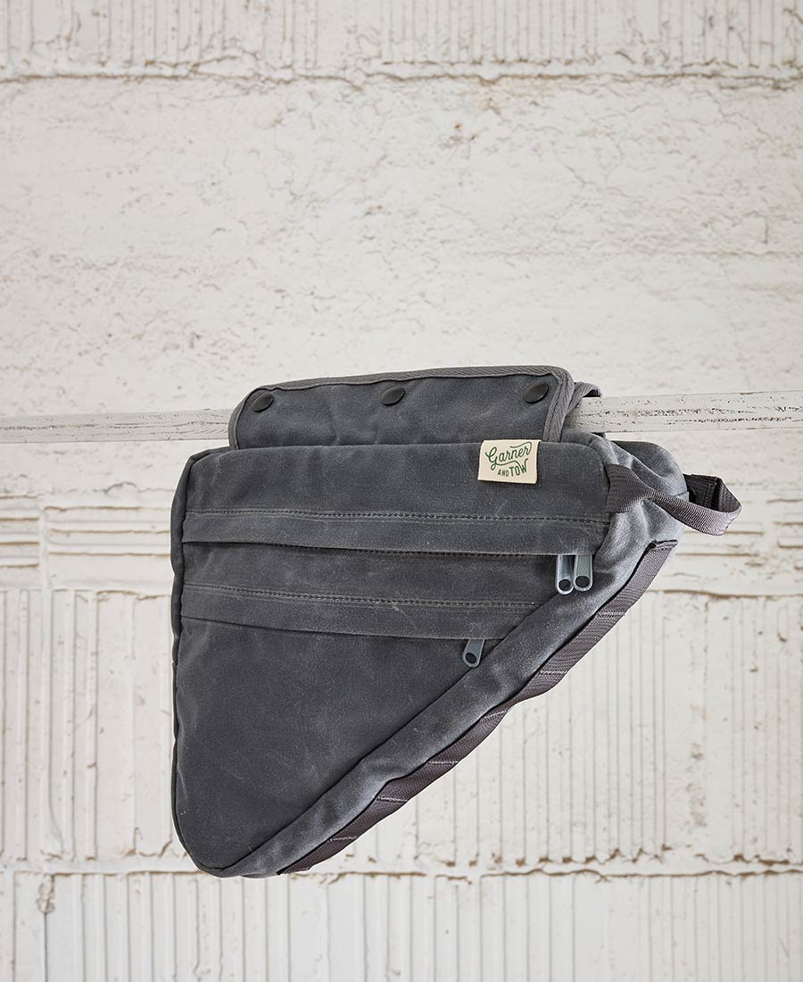 A gray bike frame bag made of waxed canvas, with snap closures and an adjustable shoulder strap, displayed against a textured background.