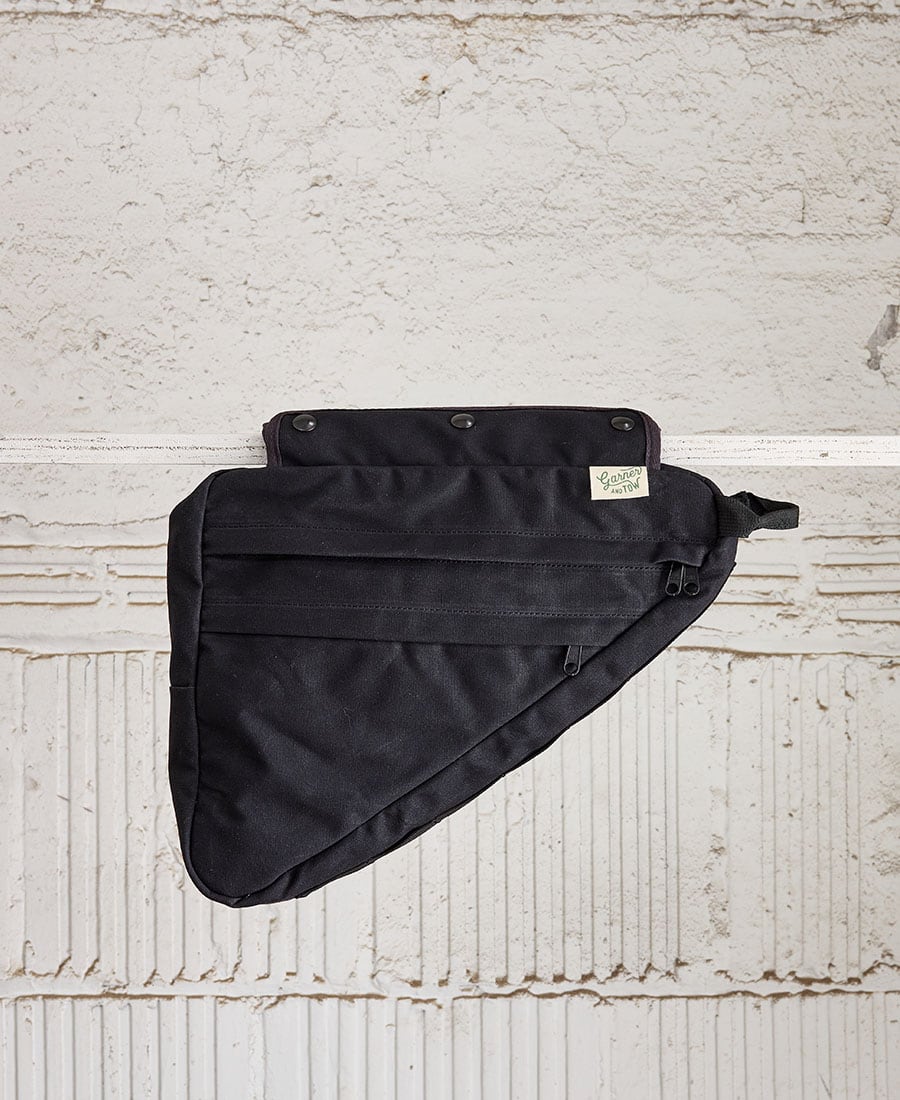 A black bike frame bag made of waxed canvas, with snap closures and an adjustable shoulder strap, displayed against a textured background.