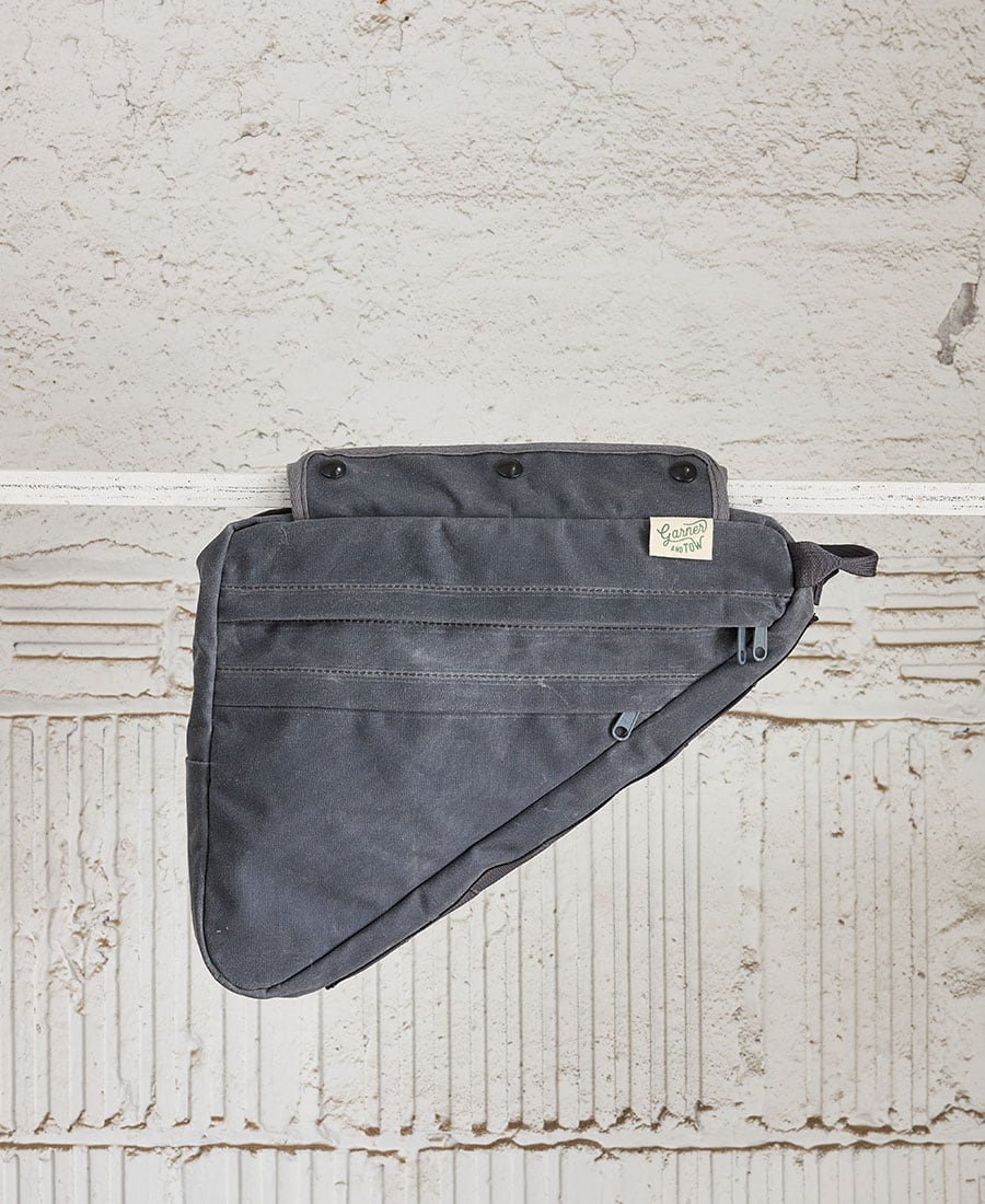 A gray frame bag designed to be attached to a bicycle's top tube, featuring multiple snap closures and zippered pockets.