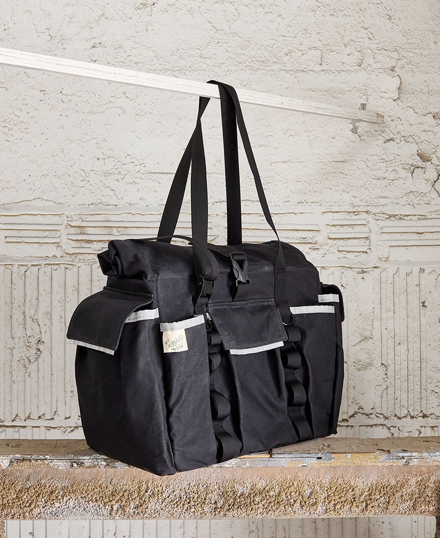 A black tote bag with multiple pockets and straps, displayed hanging on a metal rod against a concrete background.