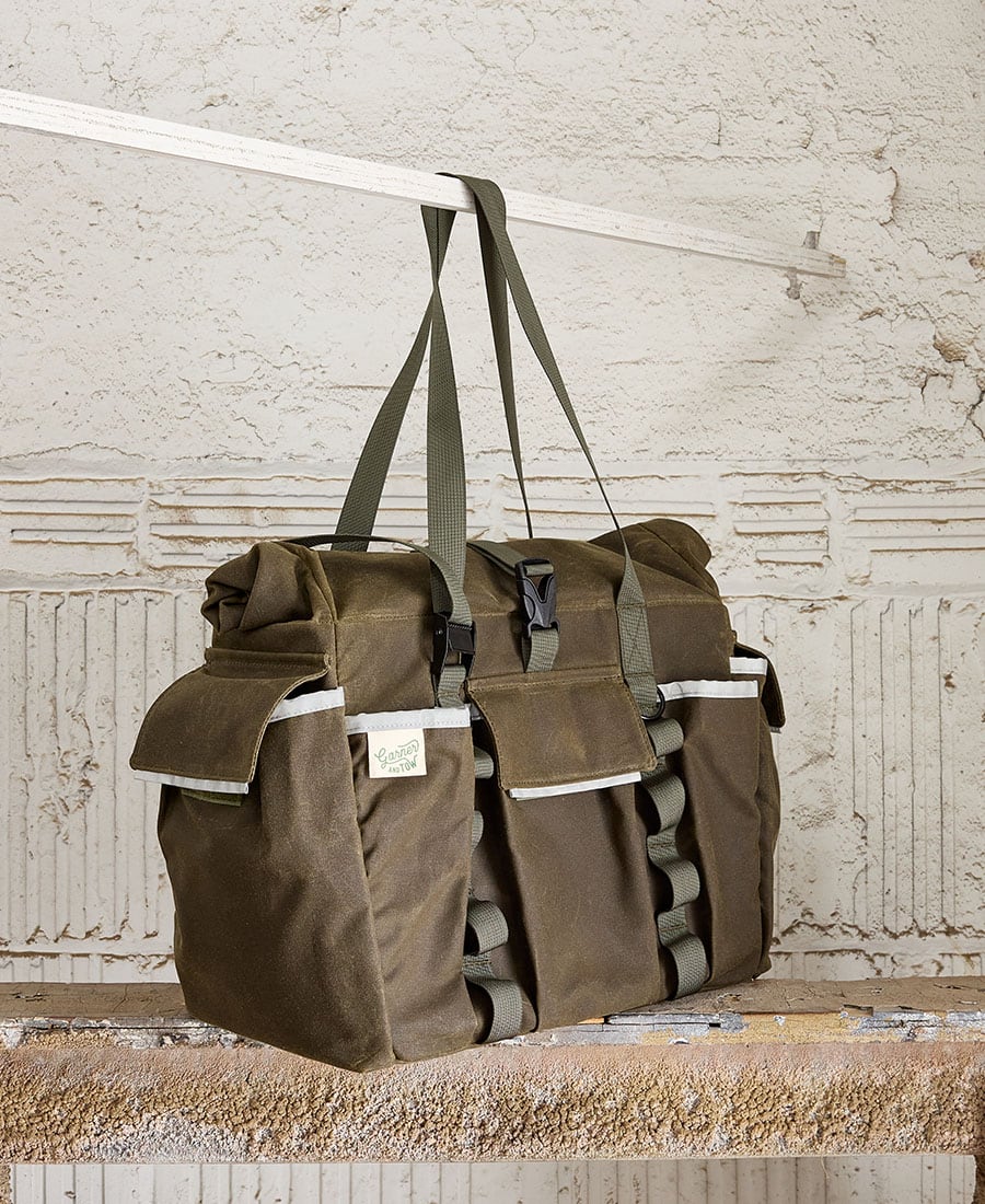 An olive tote bag with multiple pockets and straps, displayed hanging on a metal rod against a concrete background.