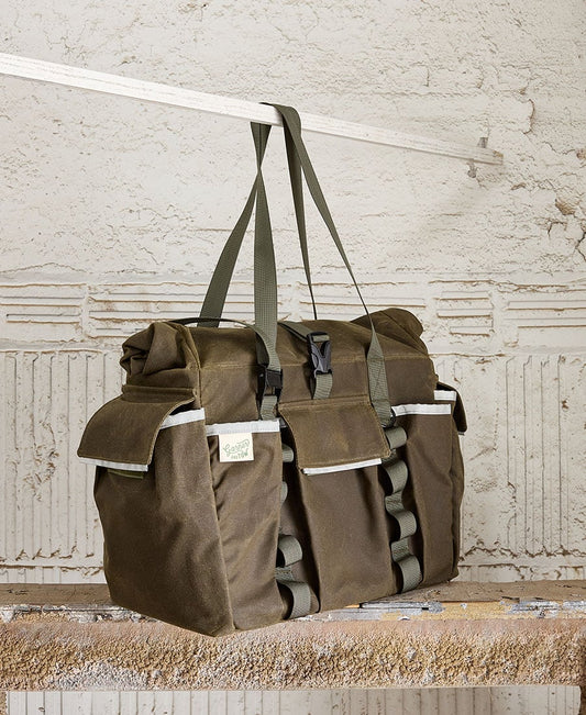 An olive tote bag with multiple pockets and straps, displayed hanging on a metal rod against a concrete background.