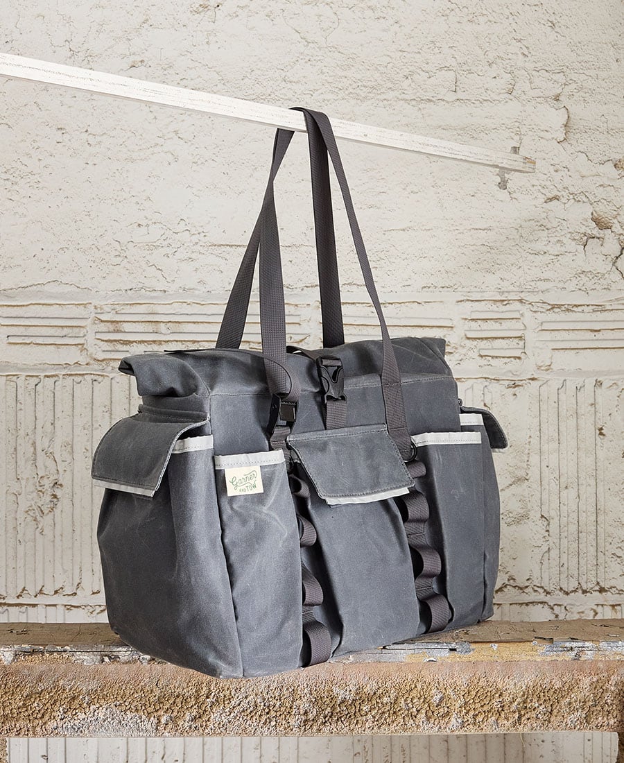 A gray tote bag with multiple external pockets and shoulder straps, displayed against an indoor backdrop.