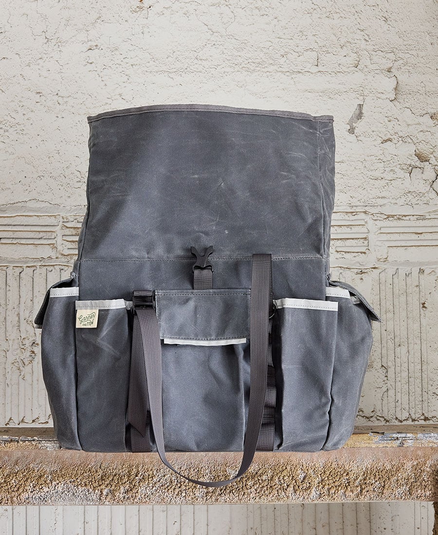 A gray tote bag shown open with multiple external pockets and shoulder straps, displayed against an indoor backdrop.