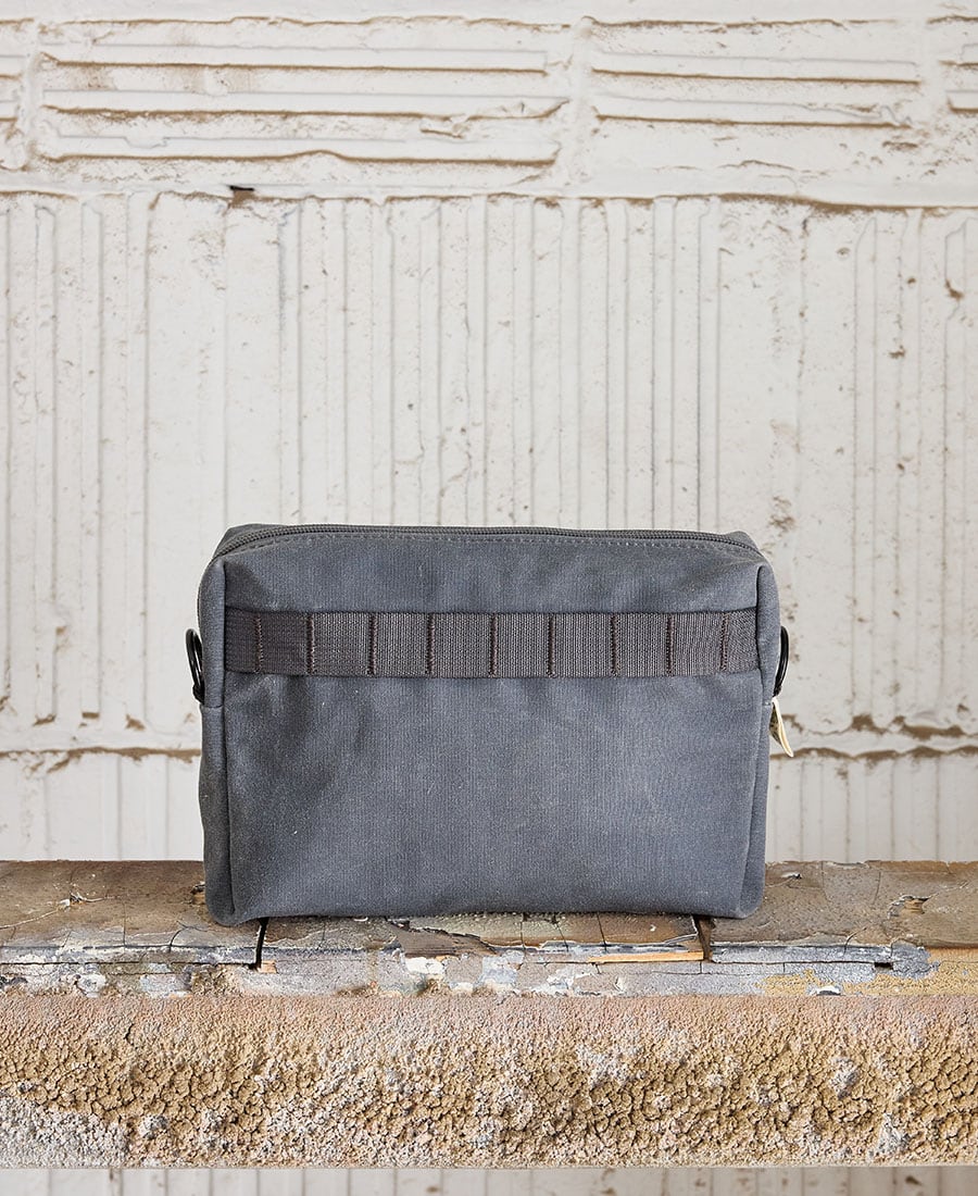 The back of a grey fabric pouch with a zippered closure, placed on a wooden surface against a white textured background.