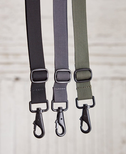 Three adjustable shoulder straps in black, gray, and green color, with metal snap hooks, displayed parallel to each other against a wooden background.