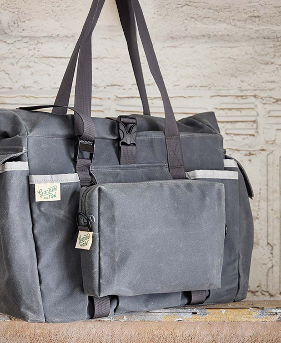 A closeup shot of a gray tote bag with multiple external pockets and shoulder straps, displayed against an indoor backdrop.